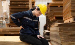 Warehouse worker sitting on the floor in front of stack of cardboard boxes with pain in the back due to frequently lifting heavy objects