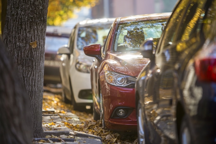 Long row of different shiny cars and vans parked along empty roadside on sunny autumn day on blurred green golden foliage bokeh background. Modern city lifestyle, vehicles parking problem concept.