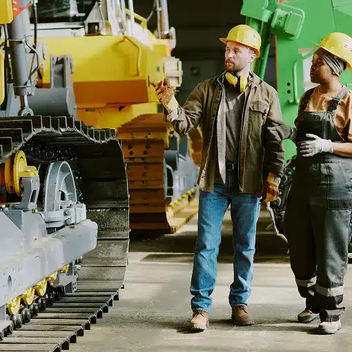 Construction workers in yellow hard hats staning in front of the tracks of large construction equipment