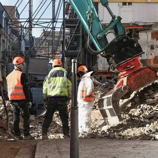 Construction workers on on a job site discussing a demolition project in front of rubble and a large crane bucket