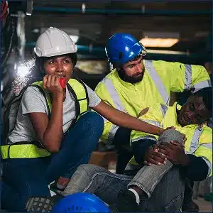 Three construction workers in yellow high visibility gear calling for aide to assist an injiured workiong on the ground holding his knee