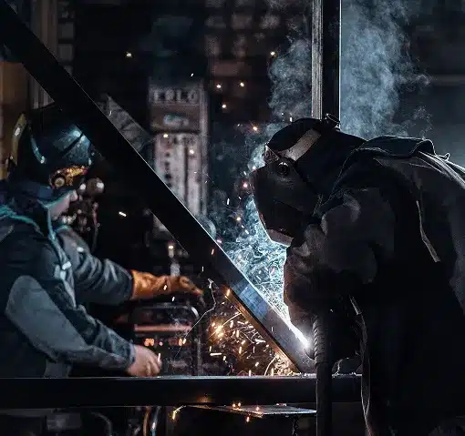 Two welders with masks on welding metal beams with a bright light from the welder throwing sparks into the air