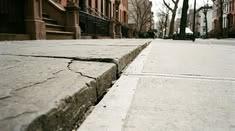 A broken and uneven sidewalk with cracked concrete slabs and exposed ground create a potential tripping hazard for New York pedestrians. 
